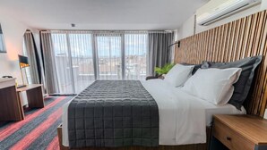 A hotel room with a bed, a desk, a chair, a potted plant, and a view of the city through large windows. - Vicolo Moda kadıköy (İstanbul)