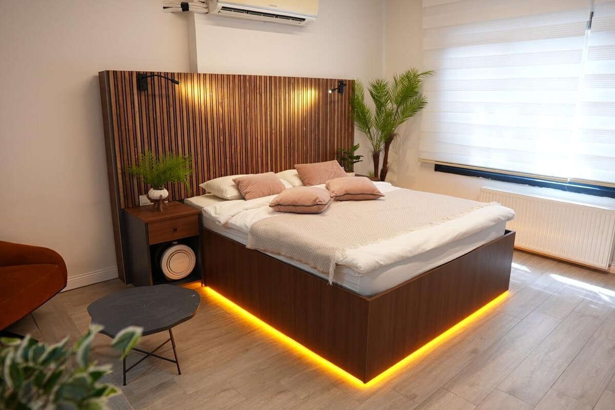 Un dormitorio moderno con cabecera de madera, una mesita de noche con lámpara, una pequeña mesa redonda y una planta en maceta.