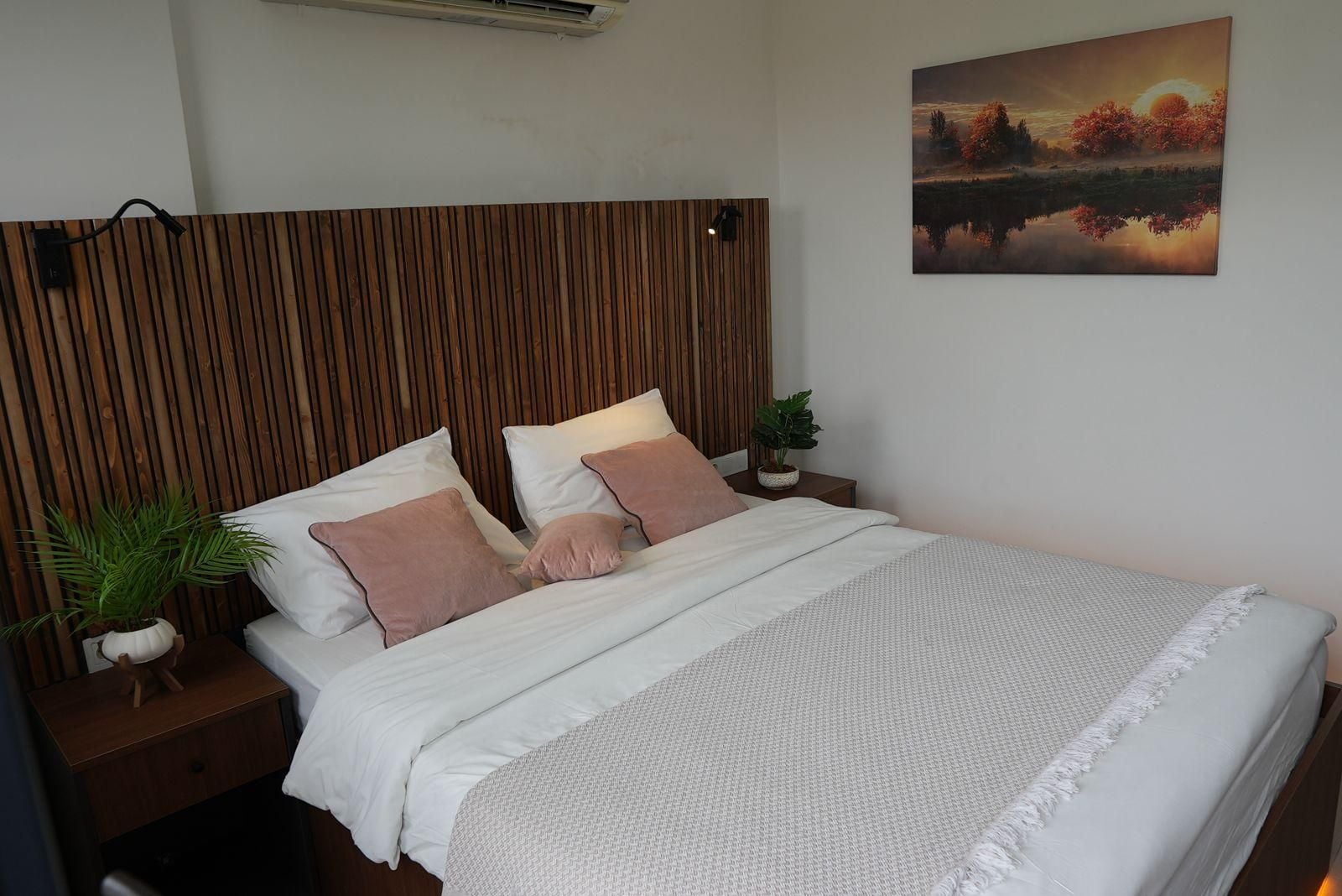 Una cama bien hecha con ropa de cama blanca y rosa, un cabecero de madera, una mesita de noche con una planta y un cuadro de un atardecer en la pared.