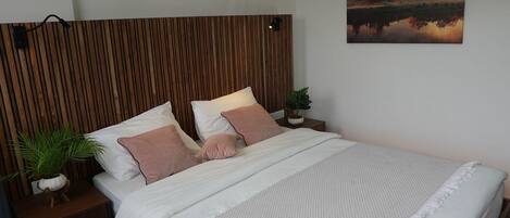 Una cama bien hecha con ropa de cama blanca y rosa, un cabecero de madera, una mesita de noche con una planta y un cuadro de un atardecer en la pared.