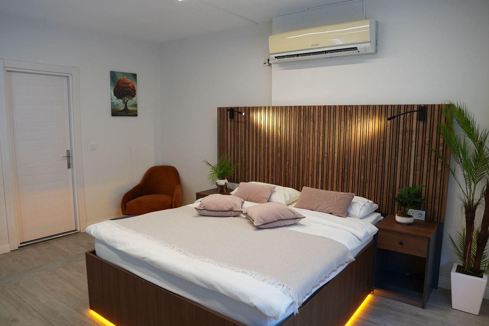 A modern bedroom with a wooden headboard, a bed with white bedding and multiple pillows, a brown chair, a bedside table with a plant, and an air conditioning unit on the wall.