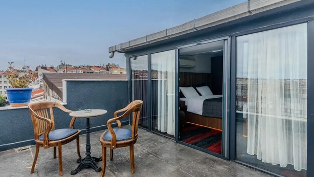 A balcony with two wooden chairs and a small round table, a sliding glass door leading to a bedroom with a bed and white curtains. - Vicolo Moda kadıköy (İstanbul)