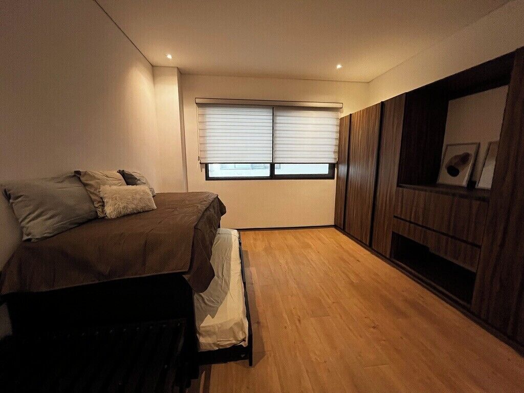 A bedroom with a bed, a window with blinds, a wooden floor, and a built-in TV shelf.
