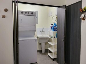 Un baño pequeño con un lavamanos blanco, un inodoro blanco, una lavadora blanca y un estante blanco con artículos de aseo.