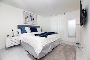 Un dormitorio moderno con cama, mesitas de noche, televisor en la pared y un amplio ventanal con cortinas.