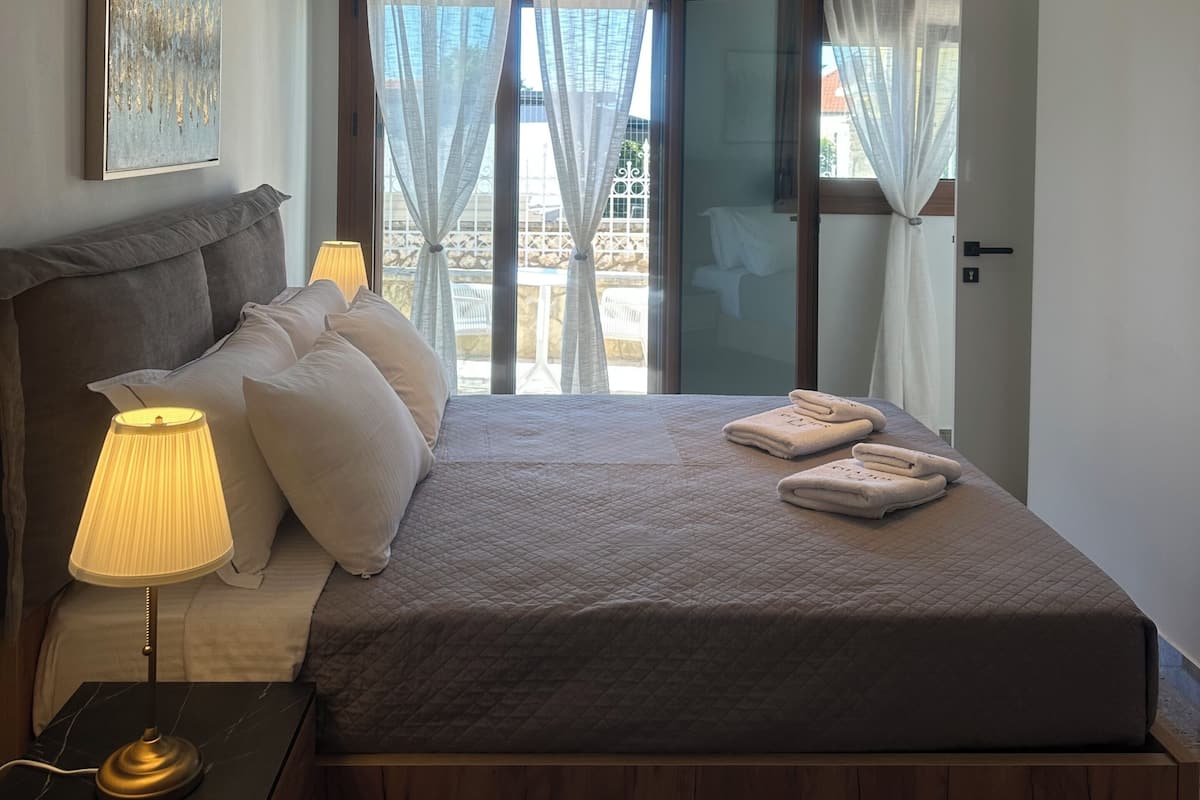A bedroom with a bed, bedside lamps, a nightstand, a wall-mounted air conditioner, and a window with curtains.
