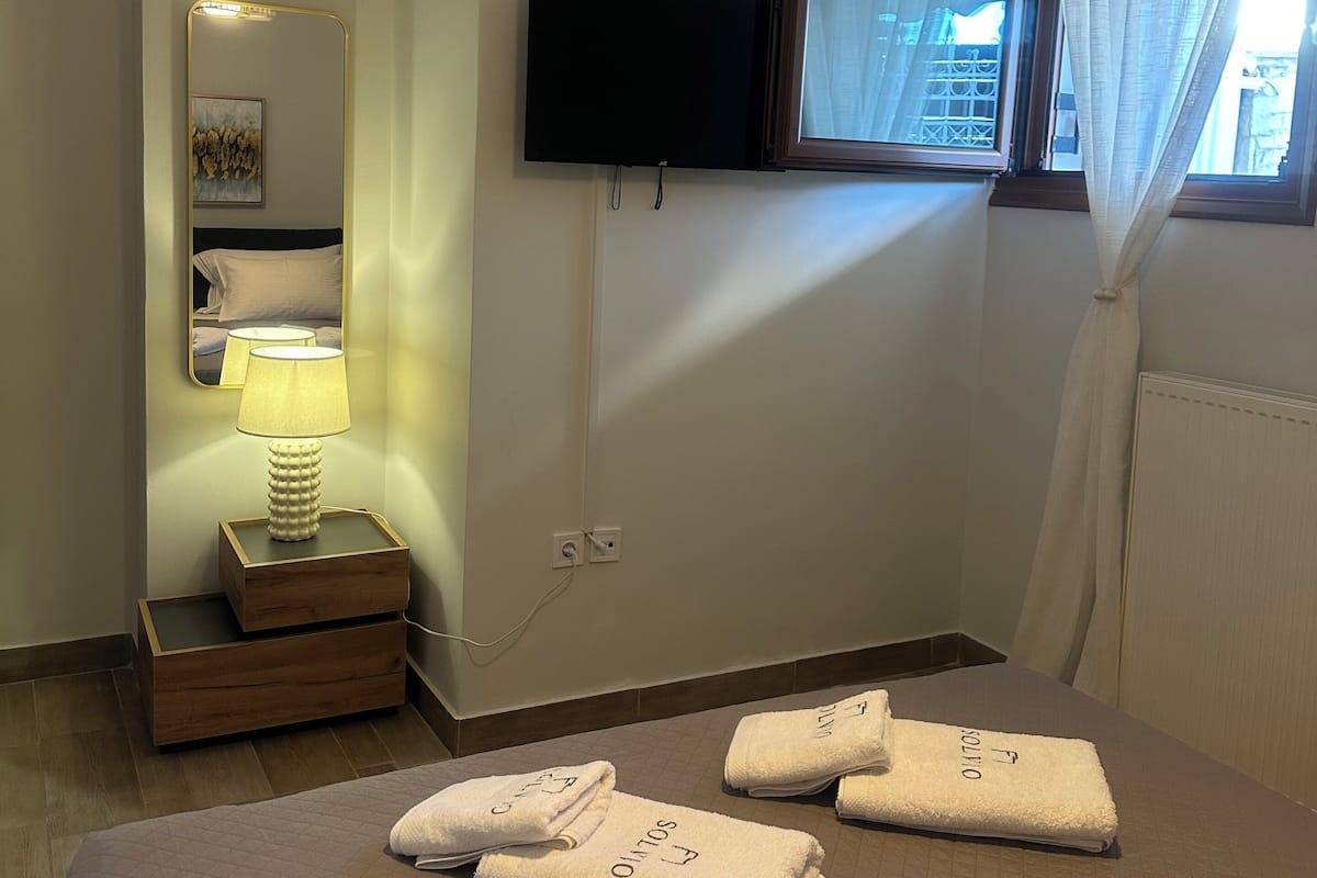 A hotel room with a bed, a TV mounted on the wall, a bedside lamp, a window with curtains, and a mirror with a light.