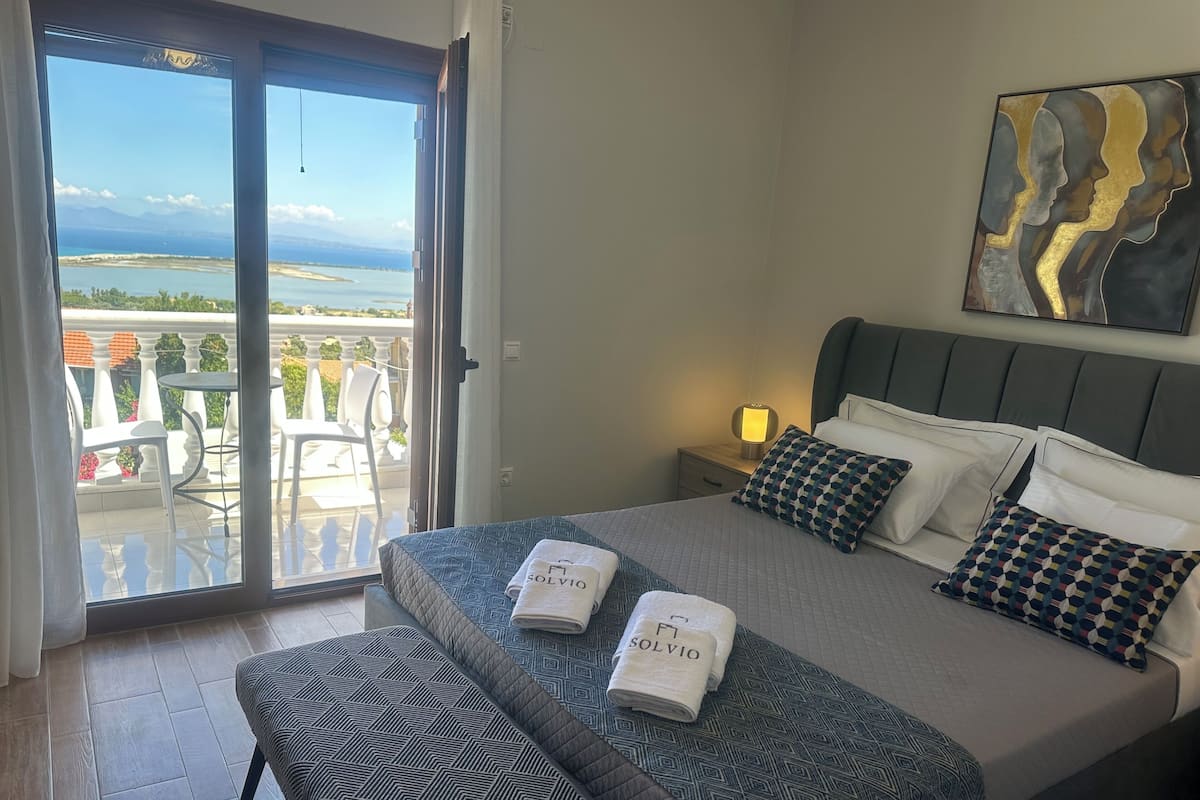 A hotel room with a bed, a bench, a nightstand, a painting, and a balcony with chairs and a table.