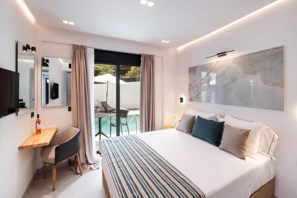 A modern hotel room with a large bed, a desk, a chair, a TV, and a balcony with a glass door.