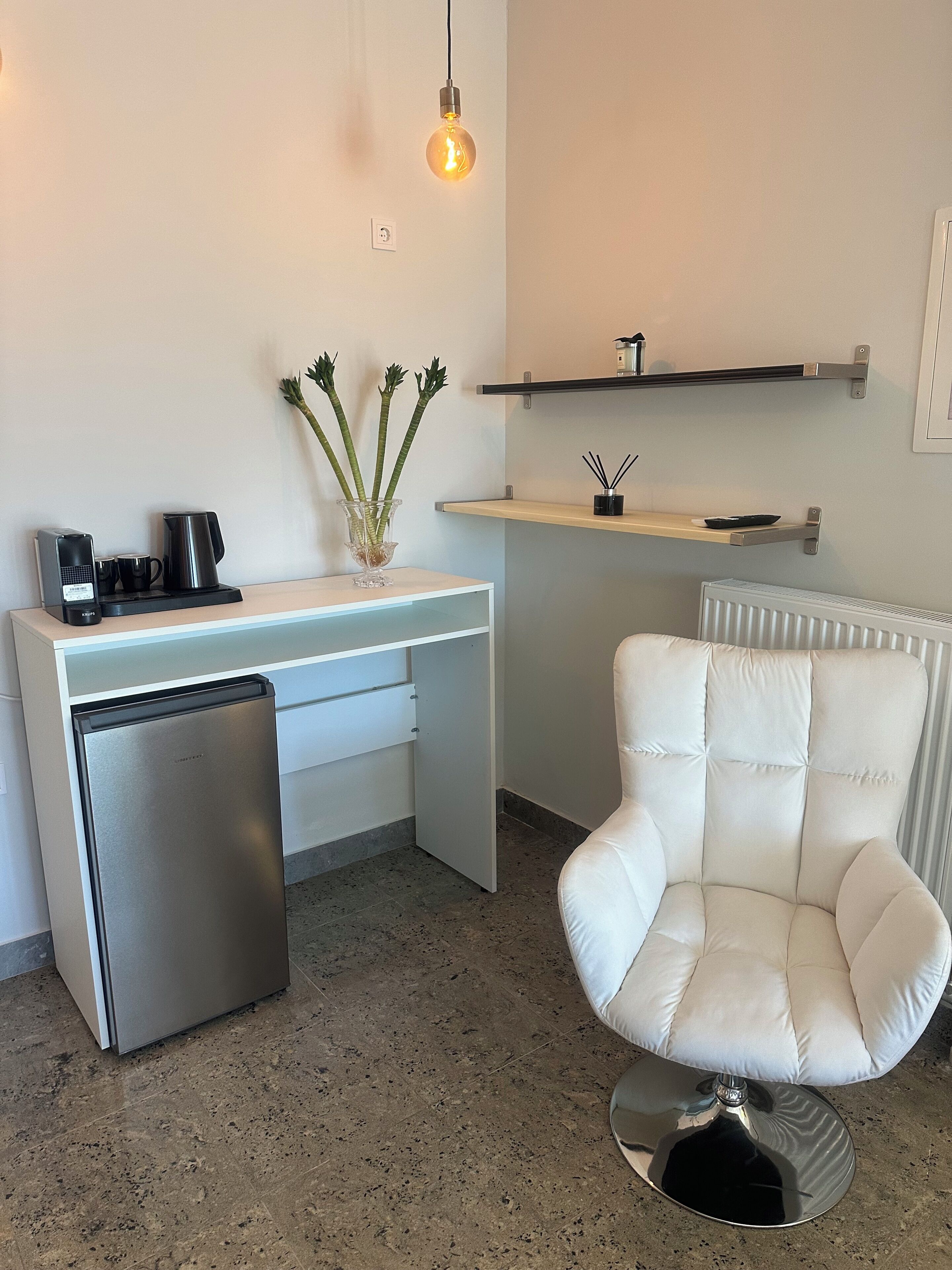 Een kitchenette met een koelkast, koffiezetapparaat en waterkoker; een witte stoel; een hangende lamp; en een radiator.