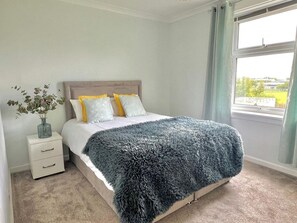 Un dormitorio con cama, mesita de noche, un jarrón con flores y una ventana con cortinas.