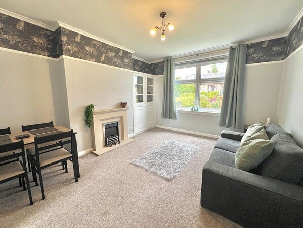 A living room with a fireplace, a sofa, a dining table with chairs, and a view through a window. - Attractive colinton property for family (Edinburgh)