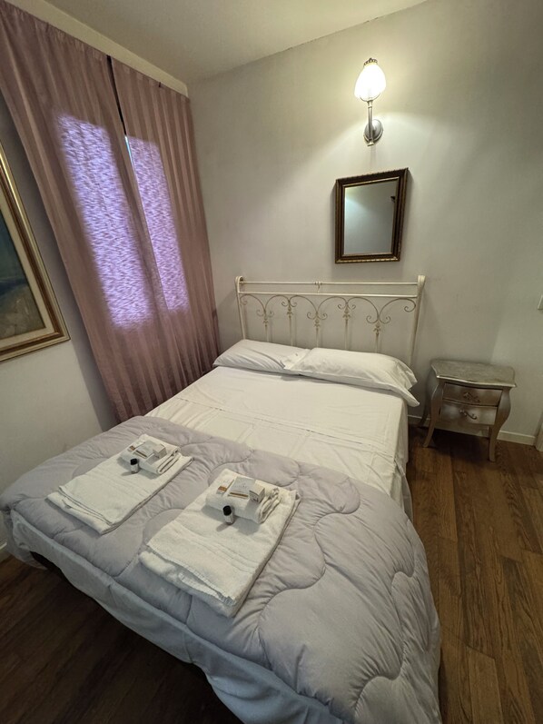 A neatly made bed with pillows and towels, a bedside table with a lamp, a window with curtains, and a framed picture on the wall. - Vrbo Property (Venezia)