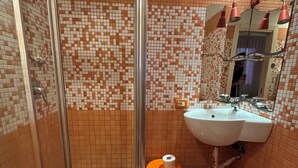 A bathroom with a shower, sink, toilet, and a small stool with a roll of toilet paper.