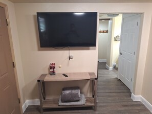 Ein Zimmer mit an der Wand montiertem Fernseher, einem kleinen Tisch mit Fernbedienung, einem Kissen und einer Steckdose.