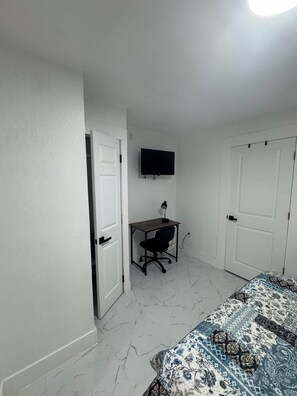 Ein Zimmer mit einem Bett, einem Schreibtisch mit Stuhl, einem an der Wand befestigten Fernseher und zwei Türen.