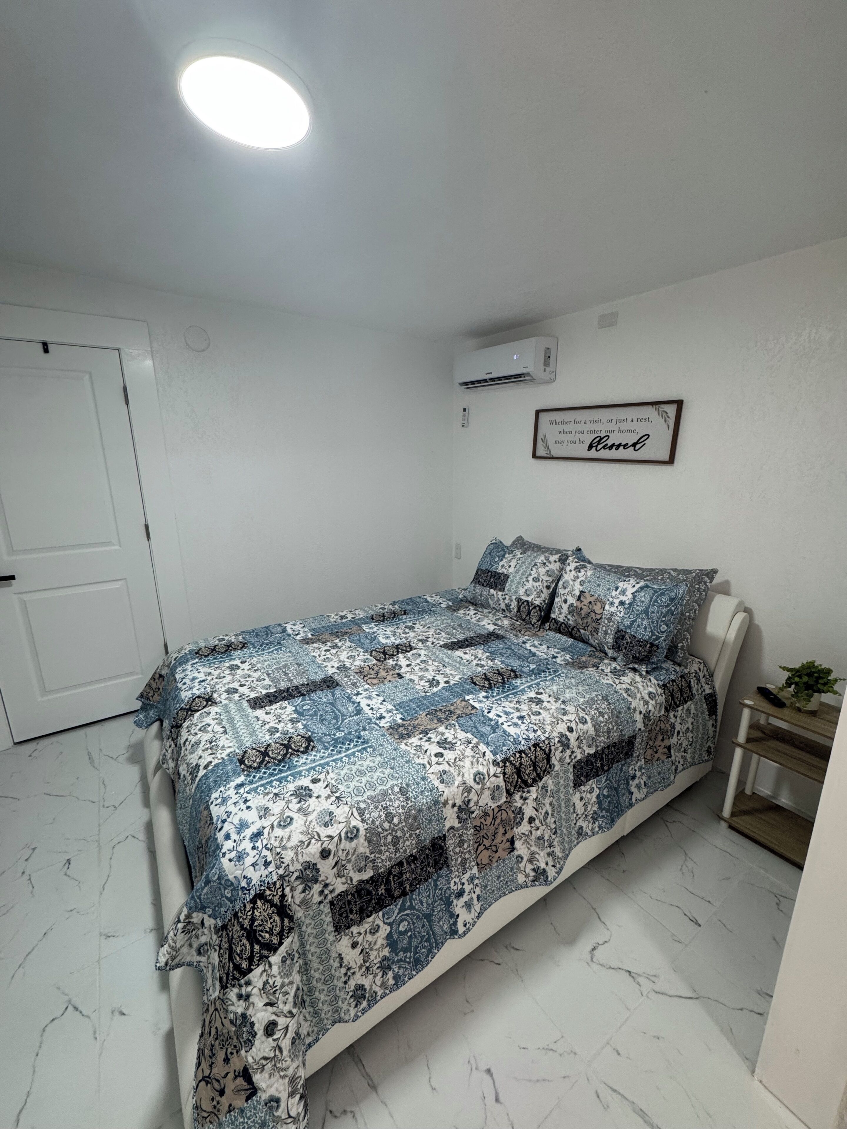A bedroom with a bed, a white door, an air conditioning unit, a framed quote on the wall, and a small plant on a nightstand.