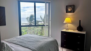 Une chambre à coucher comprenant un lit, une table de chevet, une lampe, un vase, une commode et une vue sur l’océan par une grande fenêtre.