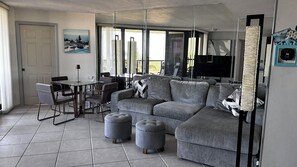 Ein modernes Wohnzimmer mit einem Glastisch, grauem Sofa, Poufs und einem großen Flachbildfernseher.