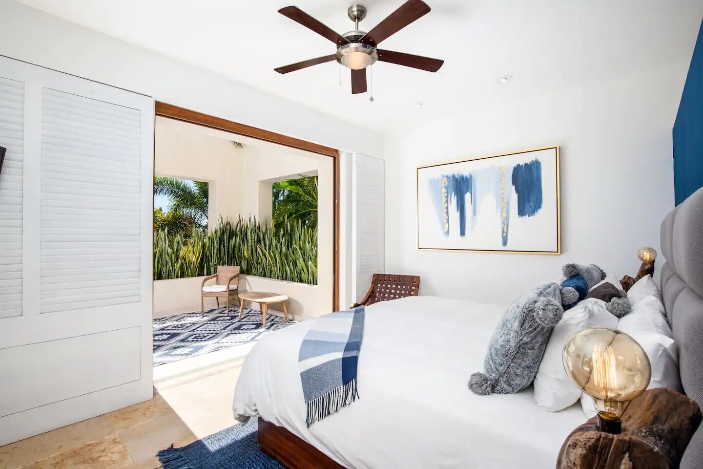 Un dormitorio con cama, ventilador de techo, ventana grande y vista a un balcón con plantas.