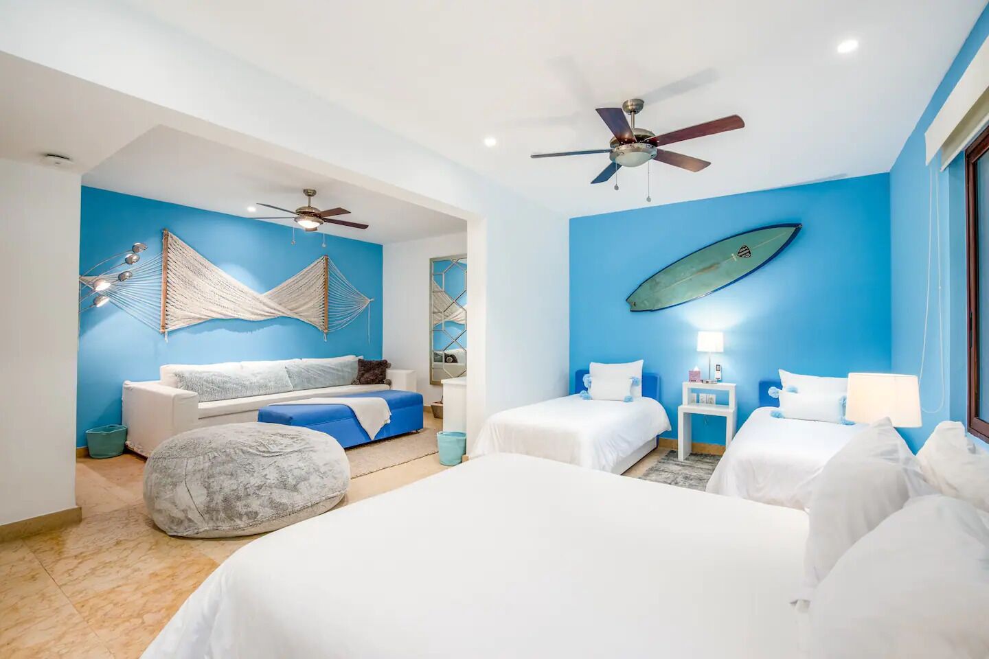 A hotel room with two beds, a sofa, a hammock, and a surfboard on the wall.