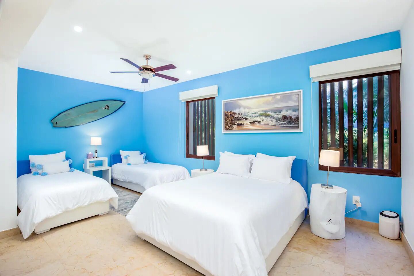 A hotel room with two beds, a ceiling fan, a surfboard decoration, a beach painting, and a window with a view of palm trees.