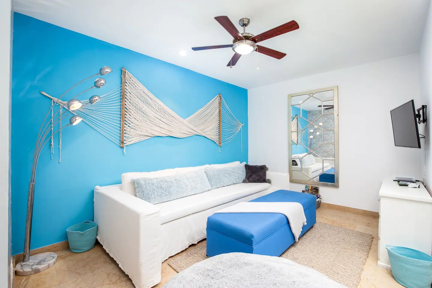 A room with a blue accent wall, a white sofa, a blue ottoman, a ceiling fan, a large mirror, and a television.