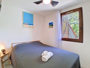 Ein Schlafzimmer mit einem Bett, einem Deckenventilator, einem Fenster mit Blick auf Grünflächen und zwei gerahmten Bildern an der Wand.