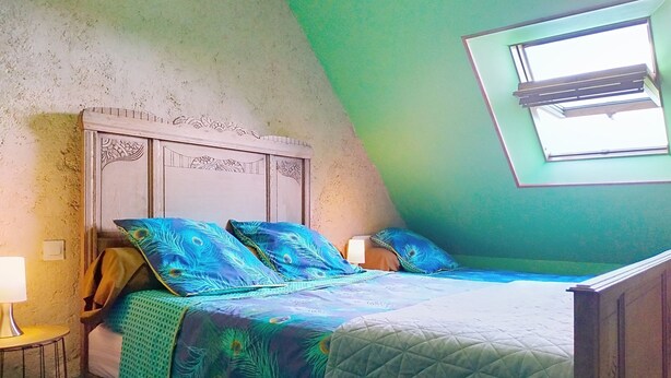 A bedroom with a sloped ceiling, a bed with peacock-patterned bedding, a wooden headboard, and a window with blinds. - L'ancienne Ferme by Interhome (Lestre)