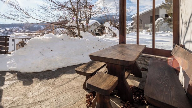 A wooden bench and table outside a building with a view of a snowy landscape and trees.