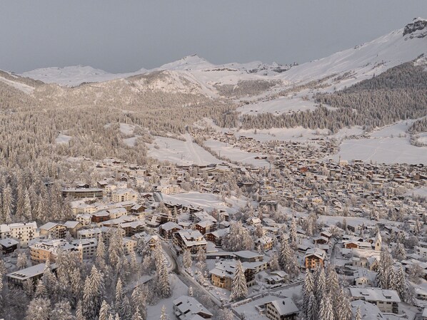 Un pueblo en la montaña cubierto de nieve, con edificios agrupados alrededor de una zona central, rodeado de laderas boscosas y montañas.