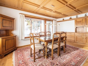 Ein traditioneller Speisesaal mit Holztisch und -stühlen, einem großen Fenster mit Vorhängen und einem gemusterten Teppich.