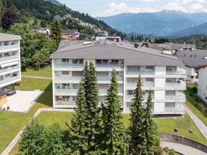 Ein mehrstöckiges Wohngebäude mit Balkonen, umgeben von Grünflächen und Bergen im Hintergrund.