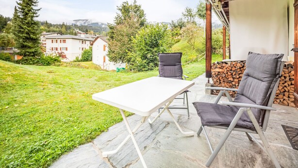 Un patio al aire libre con una mesa y sillas, techo de madera y vistas a un área verde con árboles y edificios al fondo.