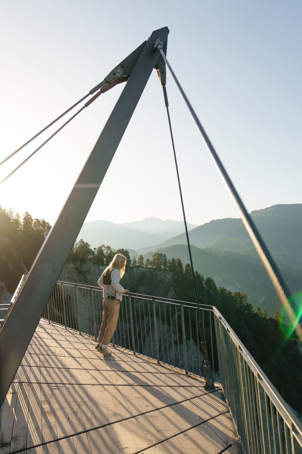 A person standing on a bridge with a camera, overlooking a mountainous landscape.
