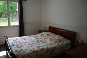 Een slaapkamer met een bed, nachtkastje, raam met gordijnen en uitzicht op groen.