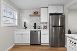 A modern kitchen with white cabinets, a stainless steel refrigerator, a dishwasher, and a window with blinds.