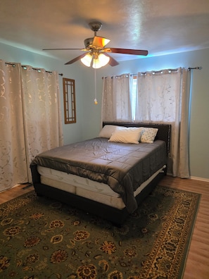 A bedroom with a bed, a ceiling fan, curtains, and a patterned rug.