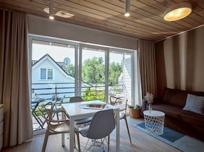 A modern living room with a dining area, large windows, and a view of a house and greenery. - Comfortable 2-room Apartments Close to the Beach (Mielno)