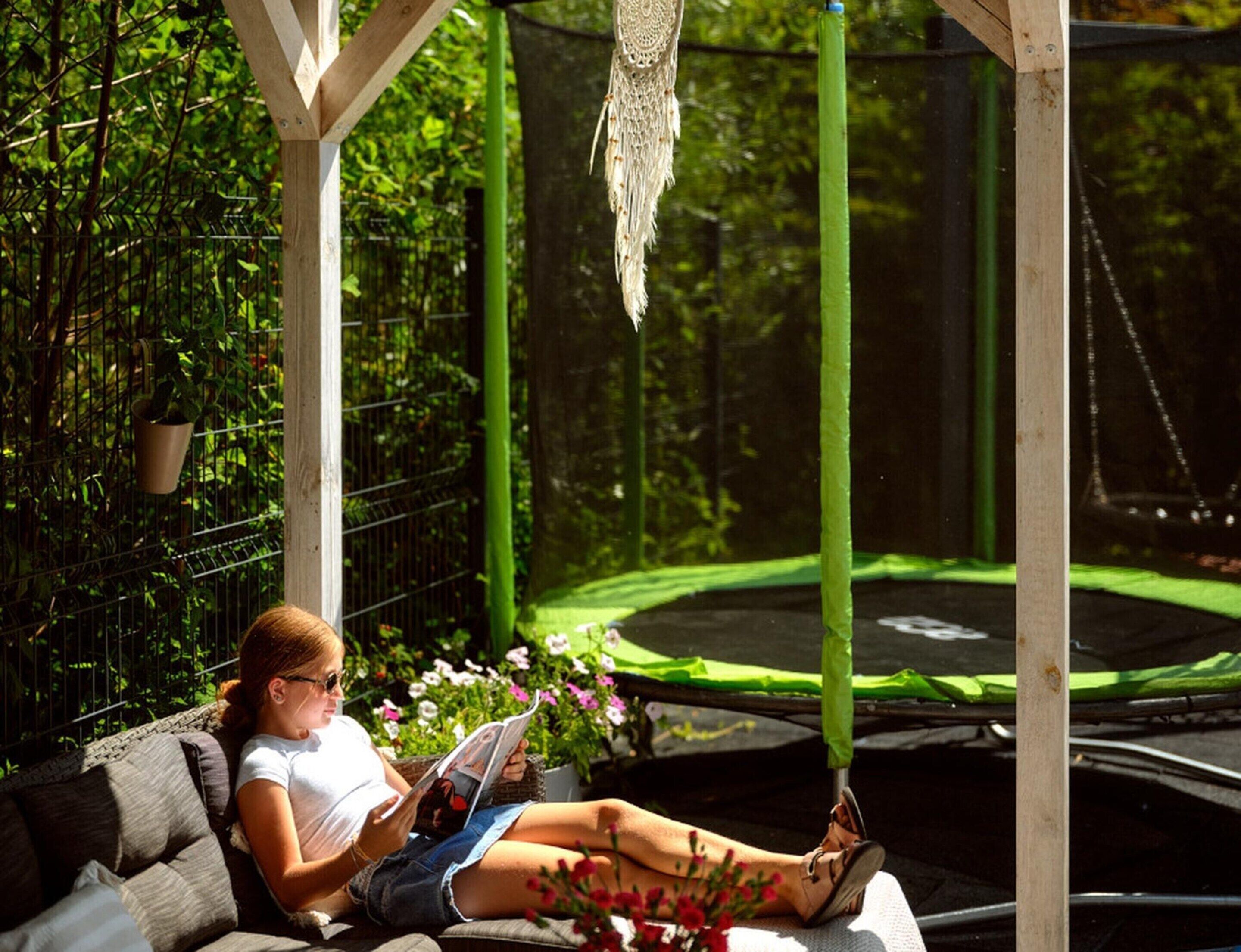 A person lounging on a cushioned bench in a garden area with a trampoline and greenery in the background.