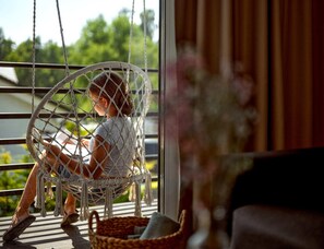 A child sitting on a white wicker swing chair by a window with a view of greenery outside. - Comfortable 2-room Apartments Close to the Beach (Mielno)