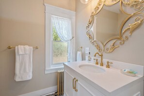 Een badkamer met een wit wastafelmeubel, gouden kranen, een grote versierde spiegel en een raam met een witte gordijn.