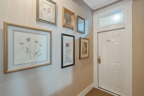 A hallway with framed botanical prints on the wall, a white door with a nameplate, and a textured carpet. - Heaton's Haven Suite B - Lovely apartment with king bed in a great location! (Baldwinsville)
