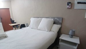 A hotel room with a bed, bedside table, lamp, and a wall-mounted thermostat.