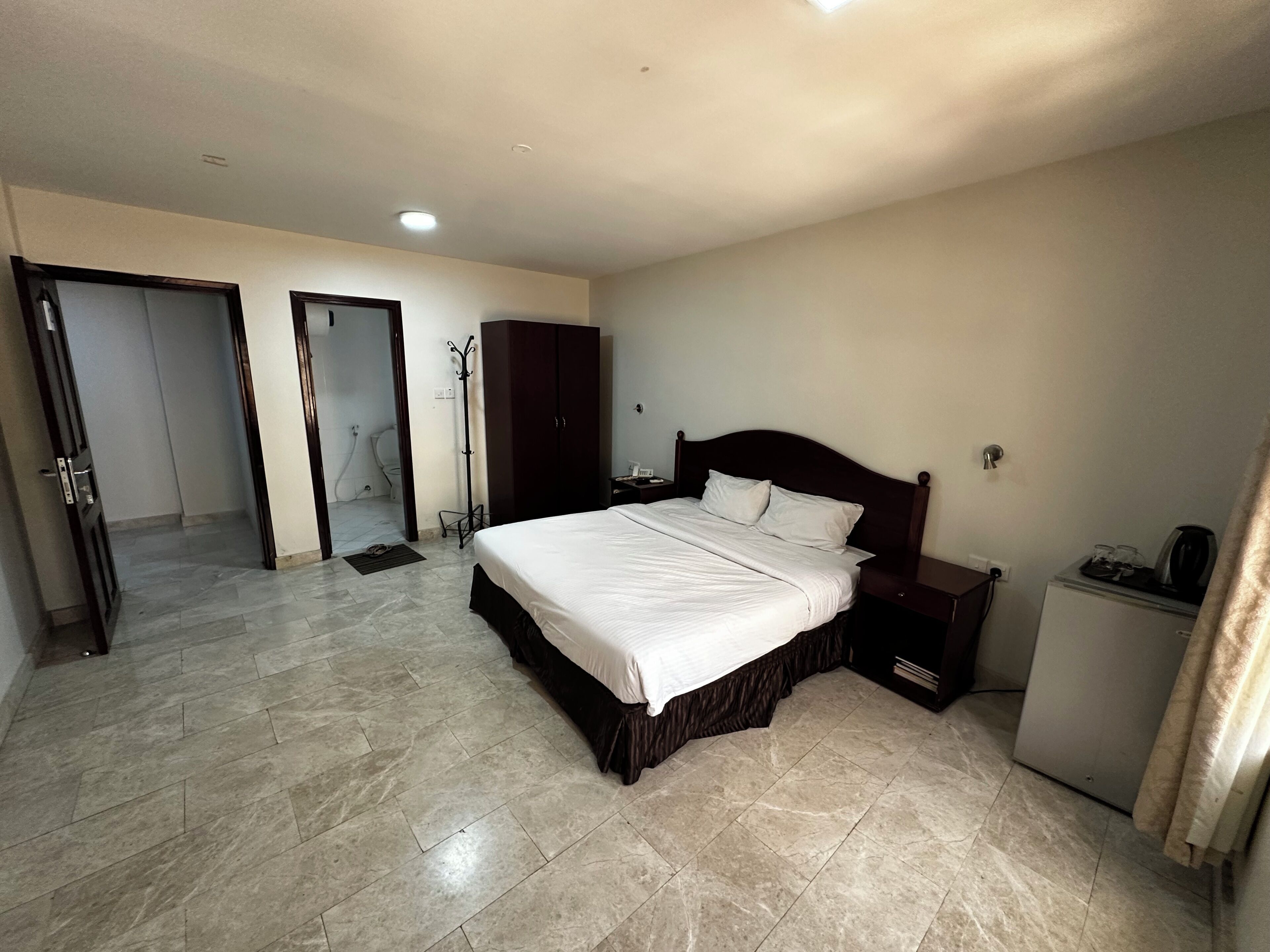 Ein Hotelzimmer mit einem großen Bett, einem Schrank, einem Schreibtisch mit Stuhl, einer kleinen Ablage und einem sichtbaren Badezimmer hinter einer offenen Tür.