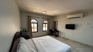 A hotel room with a bed, a desk, a chair, a television, and an air conditioning unit. - Alreef Hotel Suites (Muscat)