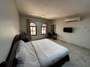 A hotel room with a bed, a desk, a chair, a television, and an air conditioning unit. - Alreef Hotel Suites (Muscat)