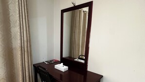 Un escritorio de madera oscura pequeño con un espejo, una silla negra, una caja de pañuelos y un cubo de basura.