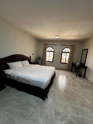 A bedroom with a bed, a desk, a chair, a window, and a mirror. - Alreef Hotel Suites (Muscat)