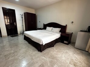 A bedroom with a large bed, a wardrobe, a desk, and a refrigerator. - Alreef Hotel Suites (Muscat)